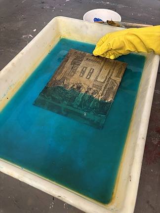 A gloved hand lifting an etched plate from a blue bath